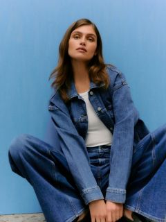 Une personne aux longs cheveux blonds lisses avec une frange porte une veste en cuir bordeaux brillante et un pantalon assorti, et s'appuie contre un mur de pierre gris clair.