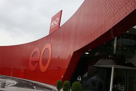Un bâtiment moderne avec une grande façade rouge incurvée portant les lettres « eo ». Des panneaux pour K&O et Giga Sport sont visibles sur le toit. L'entrée est en verre, avec des plantes en pot à côté, et une courbe passe au premier plan.