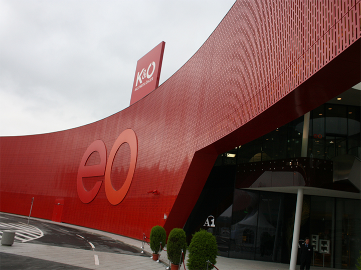 Un bâtiment moderne avec une grande façade rouge incurvée portant les lettres « eo ». Des panneaux pour K&O et Giga Sport sont visibles sur le toit. L'entrée est en verre, avec des plantes en pot à côté, et une courbe passe au premier plan.