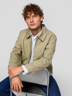 Un jeune homme portant un t-shirt noir avec l'inscription "Original Studio", un collier argenté, un bracelet argenté et une casquette de baseball rayée crème et noire se tient devant un fond gris clair uni.