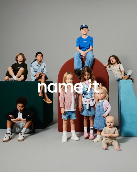 Un groupe de huit enfants différents se tient ensemble devant un fond gris, tous portant des tenues décontractées et assorties dans des tons neutres et pastel. Le texte "name it" apparaît au centre de l'image.