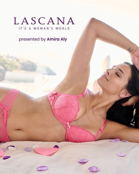 Une femme souriante en lingerie à fleurs violettes tient une petite boîte. Le texte est "LASCANA IT'S A WOMAN'S WORLD" et "LASCANA presented by Amira Aly". L'arrière-plan est doucement éclairé et décoré de manière minimaliste.
