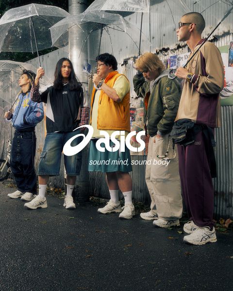 Deux personnes en tenues élégantes se tiennent avec des vélos sur une rue pavée. L'un porte une chemise bleue et un pantalon blanc, l'autre une veste et un pantalon foncés. La scène est ensoleillée et de vieux bâtiments sont visibles en arrière-plan. Le logo Asics est visible.