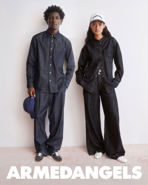 Deux jeunes adultes en vêtements en jean se tiennent ensemble en plein air, l'un avec le bras levé, l'autre avec une valise couverte d'autocollants. En bas, en lettres blanches grasses, il est écrit "ARMEDANGELS".