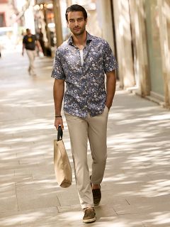 Un homme aux cheveux courts et foncés et à la barbe s'appuie sur une balustrade en pierre à l'extérieur. Il porte une veste à carreaux rouge et blanc sur un pull crème et un jean bleu. Des bâtiments urbains sont visibles en arrière-plan.