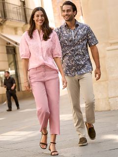 Une femme portant un manteau de fourrure à motifs et un haut rouge se tient souriante à côté d'un homme portant une veste d'hiver bleu marine et un pull beige. Ils se tiennent à l'extérieur, près d'un mur de pierre, et la femme tient un sac à main marron.
