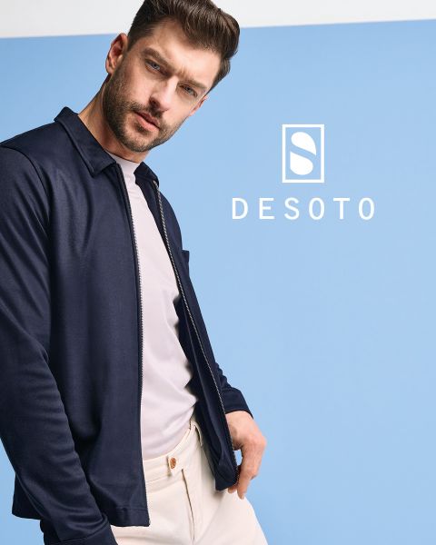 Un homme portant une chemise à manches longues à motifs et un pantalon foncé se tient devant un fond gris. Il regarde sur le côté, les mains dans les poches. L'inscription "DESOTO" et le logo DESOTO sont visibles en bas.