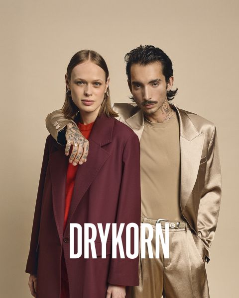 Une élégante femme âgée aux cheveux blond platine et portant un manteau de fourrure noire est assise sur un vieux canapé à côté d'un grand chien marron. Le mot "DRYKORN" est visible en bas de l'image. L'éclairage chaleureux crée une atmosphère luxueuse.