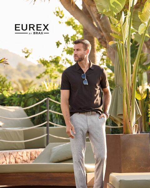 Un homme se tient à l'extérieur près de hautes herbes, portant un pull beige, une veste sans manches bleu marine et un pantalon marron, et regarde sur le côté. Le logo "EUREX by BRAX" apparaît sur le côté droit. Le ciel est nuageux.