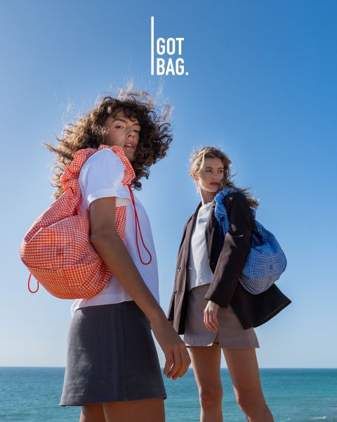 Un homme portant des lunettes de soleil tient de grands sacs gris devant un fond à rayures foncées. En dessous de lui, différents sacs de différentes tailles et couleurs sont soigneusement exposés. Le logo en haut à droite est « GOT BAG ».