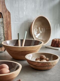 Sur une table de cuisine, il y a trois saladiers en bois de différentes tailles. Dans un saladier, il y a des noix, dans un autre, deux œufs, et dans le plus grand, des ustensiles. À l'arrière-plan, on peut voir une râpe, une planche à découper et un saladier fixé au mur.