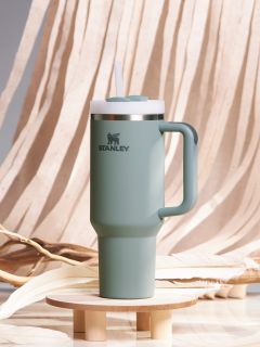 Une gourde de voyage Stanley verte avec une anse et un couvercle à paille est posée sur un petit socle en bois. L'arrière-plan est composé d'un tissu beige fluide et de branches décoratives.