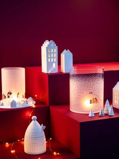 Une exposition festive avec des maisons en céramique lumineuses, des bougeoirs en verre opale avec des motifs hivernaux et de petits arbres blancs, tous placés sur des socles rouges avec des guirlandes lumineuses.