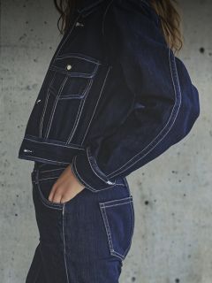 Une femme avec un bonnet blanc, un t-shirt blanc et un jean bleu pose avec le bras tendu dans une pièce blanche et moderne avec des fleurs au-dessus.