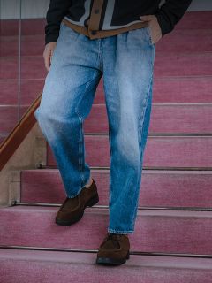 Un homme en t-shirt blanc, veste en jean foncée et jean se tient à une balustrade appuyée contre un mur en béton et une clôture, surmontée d'un auvent jaune.
