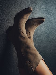 Gros plan sur les jambes d'une personne portant des bottes de cowboy taupe en daim avec des coutures décoratives, appuyées contre un mur texturé gris foncé.