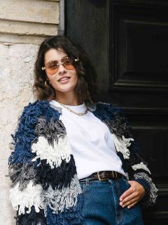 Une femme aux cheveux bruns courts, portant des lunettes de soleil, un cardigan épais bleu et blanc, un t-shirt blanc et un jean bleu, se tient nonchalamment devant un mur de pierre et une porte sombre, et sourit avec assurance.