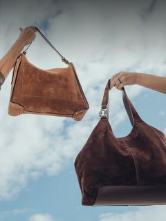 Deux mains tiennent des sacs à main en daim devant un ciel bleu nuageux ; un sac est brun clair, l'autre brun foncé.