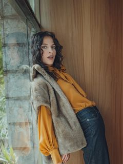 Une femme aux cheveux ondulés et foncés est appuyée contre un mur en bois près d'une fenêtre. Elle porte un chemisier jaune à manches bouffantes, un gilet en fourrure marron et un jean foncé. Son expression est détendue alors qu'elle regarde l'appareil photo.