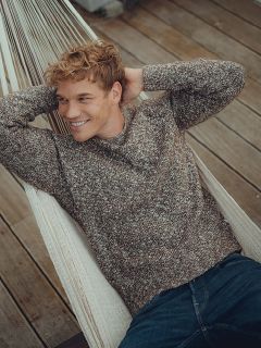 Une personne souriante aux cheveux bouclés, portant un pull en tricot marron et un jean bleu, est allongée dans un hamac blanc sur une terrasse en bois, les mains derrière la tête.