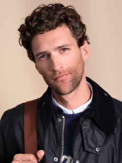 Un homme aux cheveux bruns bouclés et aux yeux bleus pose devant un fond neutre. Il porte une veste foncée sur une chemise à col et un pull, et a une bande marron sur l'épaule.