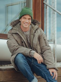 Un homme souriant, portant un bonnet en tricot vert, un manteau d'hiver gris, un jean bleu et un pull à motifs, est assis sur un escalier en bois à l'extérieur, à côté d'une balustrade en métal.