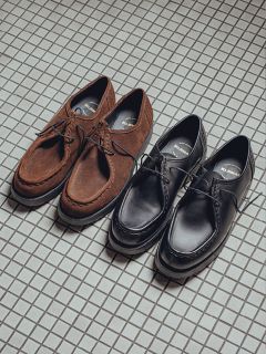 Deux paires de chaussures à lacets sont côte à côte sur un sol carrelé : une paire en daim marron et l'autre en cuir noir. Les deux chaussures ont un design similaire de style mocassin.
