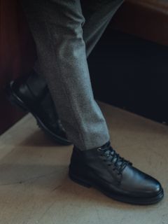 Personne en pantalon gris et bottes à lacets en cuir noir, une jambe croisée sur l'autre, se tient à l'intérieur sur un sol clair.