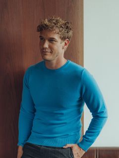 Un homme aux cheveux blonds courts et bouclés, portant un pull bleu clair et un pantalon foncé, se tient à l'intérieur devant un mur en bois et blanc et regarde légèrement sur le côté avec une expression détendue.