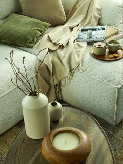 Scène de salon confortable avec un canapé doux et clair, des coussins verts et beiges, un jeté beige, un plateau avec du thé et des biscuits, un magazine, des livres et une table basse en bois avec un vase, une bougie et des branches décoratives.