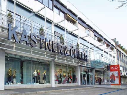 Das Bild zeigt die Ladenfront von Kastner & Öhler mit einem großen rot-weißen Logo auf der linken Seite und einer modernen Glasfassade. In den Fenstern unter dem markanten Namensschild des Ladens stehen Schaufensterpuppen in stilvoller Kleidung.
