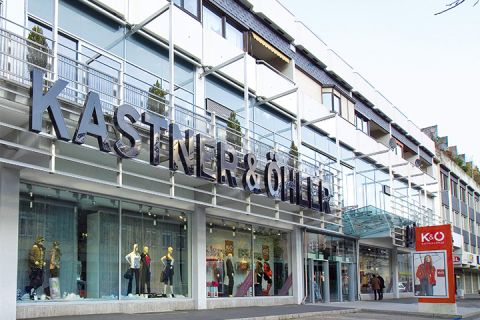 L'image montre la façade du magasin Kastner & Öhler avec un grand logo rouge et blanc sur le côté gauche et une façade en verre moderne. Dans les fenêtres, sous le nom distinctif du magasin, se trouvent des mannequins portant des vêtements élégants.
