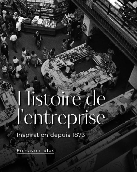 Schwarz-Weiß-Foto einer belebten Markthalle oder eines Kaufhauses mit Menschen, die an verschiedenen Schaltern einkaufen. Der überlagerte Text lautet: "Histoire de l'entreprise Inspiration depuis 1873.