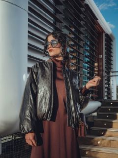 Une femme avec de grandes lunettes de soleil et une veste en cuir sombre est appuyée contre un mur, les mains croisées derrière la tête, et pose avec assurance au soleil.
