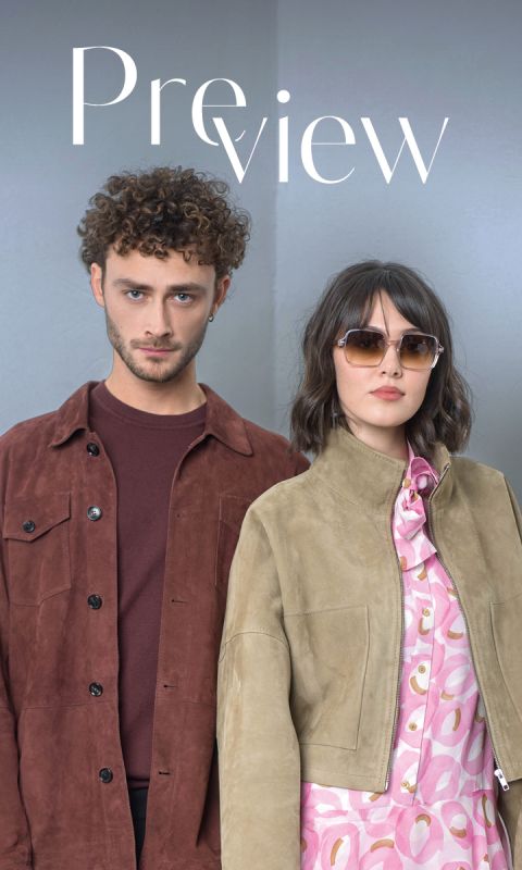 Un homme portant une veste marron et une femme portant des lunettes de soleil et une veste marron clair sur une robe à motifs roses se tiennent côte à côte devant un fond clair et uni.