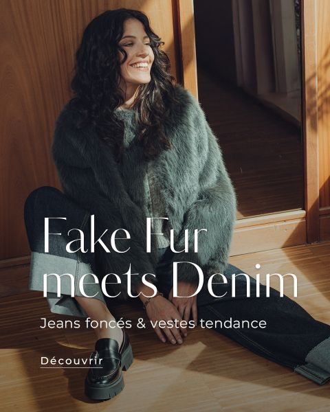 Une femme portant une veste en fausse fourrure marron clair et un jean foncé est assise avec assurance devant un fond vert. Le texte superposé indique : "La fausse fourrure rencontre le denim. Jeans foncés & vestes tendance. Découvrir.".