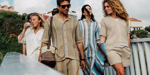 Un groupe de cinq personnes prend un selfie à l'extérieur, près de l'eau, avec des arbres et un coucher de soleil en arrière-plan. Ils sourient et posent de manière ludique, vêtus de tenues décontractées. Au-dessus d'eux, on peut voir le texte « KnowledgeCotton Apparel » et un logo de hibou.