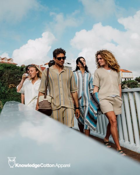 Un groupe de cinq personnes prend un selfie à l'extérieur, près de l'eau, avec des arbres et un coucher de soleil en arrière-plan. Ils sourient et posent de manière ludique, vêtus de tenues décontractées. Au-dessus d'eux, on peut voir le texte « KnowledgeCotton Apparel » et un logo de hibou.