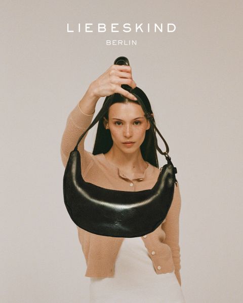 Une femme souriante, vêtue d'une chemise noire à manches longues et d'un jean, porte un sac marron clair en bandoulière. Le texte "Liebeskind Berlin" apparaît en bas de l'image.