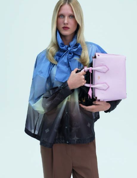 Un sac à main de créateur vert avec un motif monogramme discret repose sur une surface grise, avec un objet géométrique bleu au premier plan et une ombre portée à l'arrière-plan.