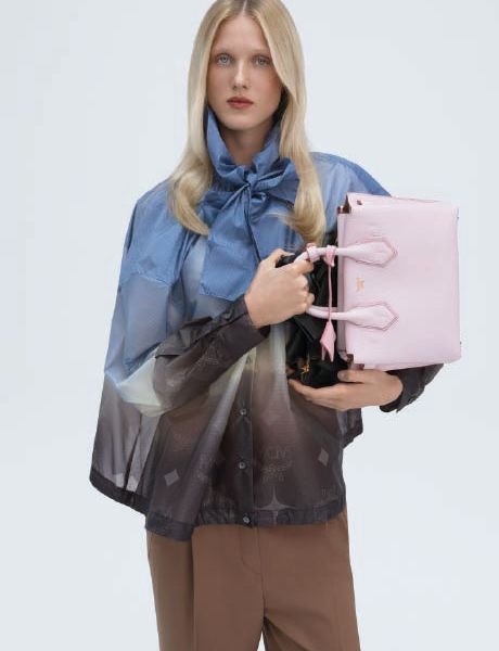 Un sac à main de créateur vert avec un motif monogramme discret repose sur une surface grise, avec un objet géométrique bleu au premier plan et une ombre portée à l'arrière-plan.