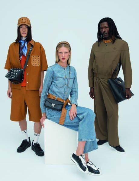 Un sac de créateur MCM bleu avec un motif de logo blanc est posé sur des marches en bois ornées de porte-clés colorés représentant des animaux et des objets, notamment une fusée, un lapin et un ours.