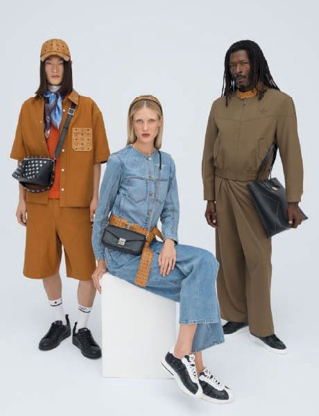 Un sac de créateur MCM bleu avec un motif de logo blanc est posé sur des marches en bois ornées de porte-clés colorés représentant des animaux et des objets, notamment une fusée, un lapin et un ours.