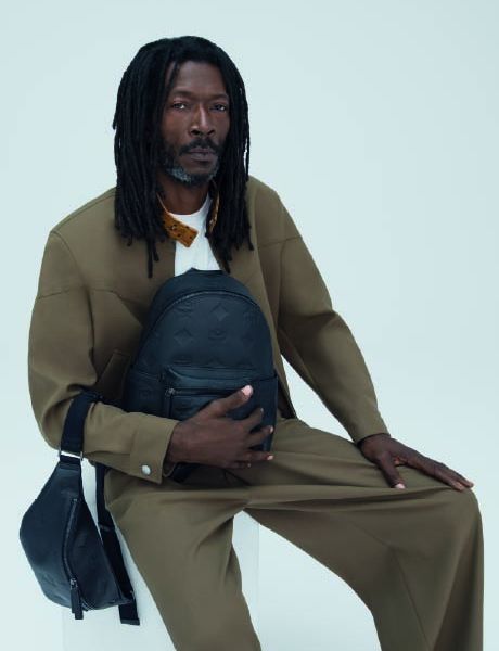 Un sac à dos de créateur vert et un sac à bandoulière assorti sont posés sur une pile de boîtes orange et marron devant un mur texturé beige et un sol carrelé.