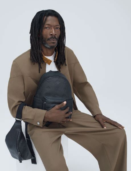 Un sac à dos de créateur vert et un sac à bandoulière assorti sont posés sur une pile de boîtes orange et marron devant un mur texturé beige et un sol carrelé.