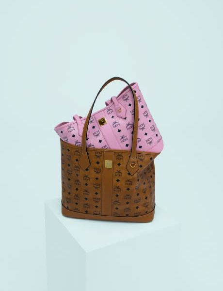 Une femme portant une veste en peau de mouton retournée marron clair, un short noir et des bottes se tient sur une marche en pierre et tient un grand sac. En arrière-plan, un homme en vêtements colorés semble flotter horizontalement au-dessus des marches. Derrière eux, on peut voir des arbres.