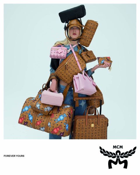 Trois femmes portant des manteaux marron clair et des chapeaux orange traversent une rue, portant des sacs MCM bleus avec un logo MCM blanc au premier plan. L'image a un effet miroir qui donne l'impression que la scène est dupliquée.