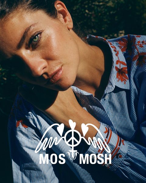 Une femme aux longs cheveux blonds ondulés porte une chemise verte à motifs blancs. Elle regarde par-dessus son épaule et se tient devant un bâtiment moderne. Le logo "MOS MOSH" se trouve dans le coin supérieur gauche.