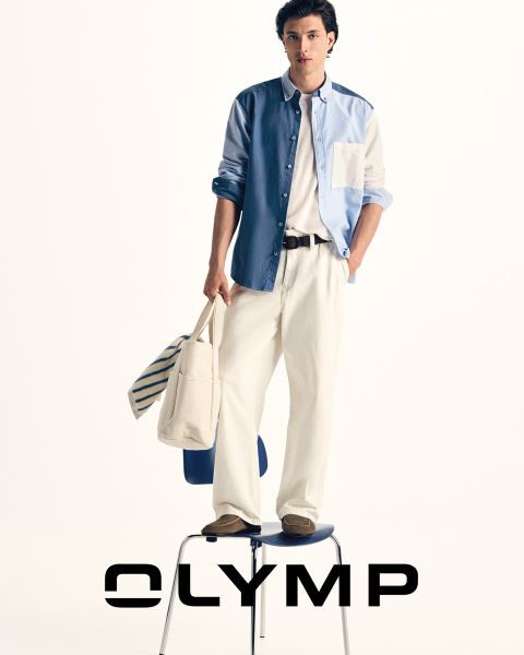 Un homme en pantalon de costume, chemise à carreaux et cravate, avec un pull drapé sur les épaules, se tient devant un fond bleu foncé, à côté duquel on peut lire le mot "OLYMP".