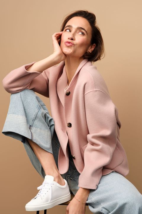 Une femme en cardigan rose et en jean est assise de manière ludique sur un tabouret, le menton appuyé sur le genou. Elle penche la tête et fait une moue espiègle, la main posée sur sa joue. Elle porte des baskets blanches et pose devant un fond beige.